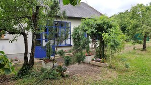 A house with a blue door and windows, surrounded by a garden with various plants and trees. - Maison de Vacance Tout Confort Avec Piscine (saint benoit du sault)