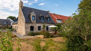 Een stenen huis met een schuin dak, schoorsteen en een kleine tuin met gras en struiken.