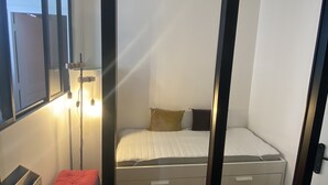 A small, single-bed room with a window, a bedside table, and a red ottoman.