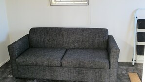 A room with a grey sofa, a window with blinds, a ladder, and a tiled floor.