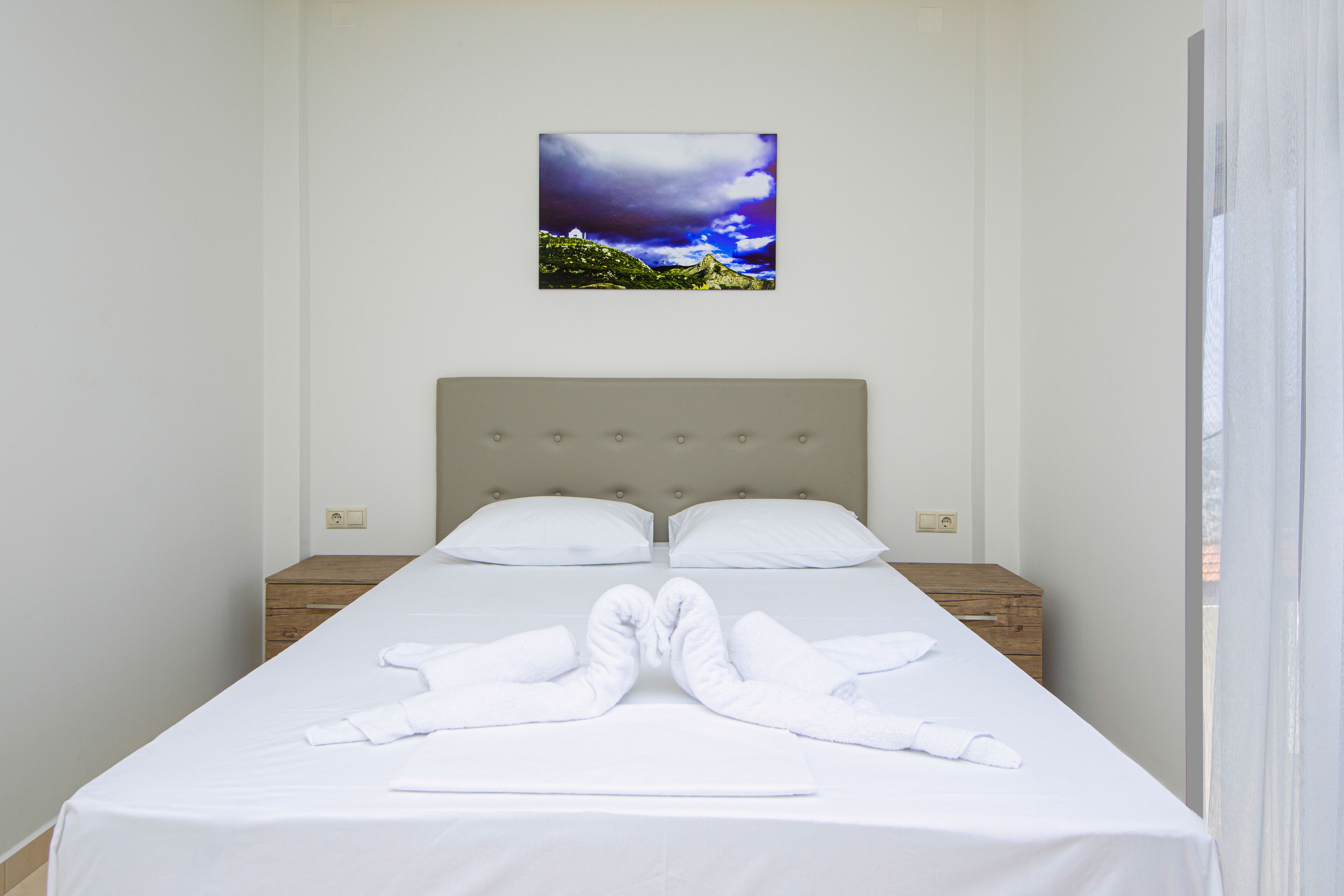 A neatly made bed with white linens and towels arranged in a heart shape, a wooden nightstand, and a framed picture of a landscape on the wall.