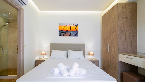 A neatly made bed with rolled towels, a wooden wardrobe, a bedside table with a lamp, and a glass shower enclosure. - Amara Suites and Studios (Hersonissos)