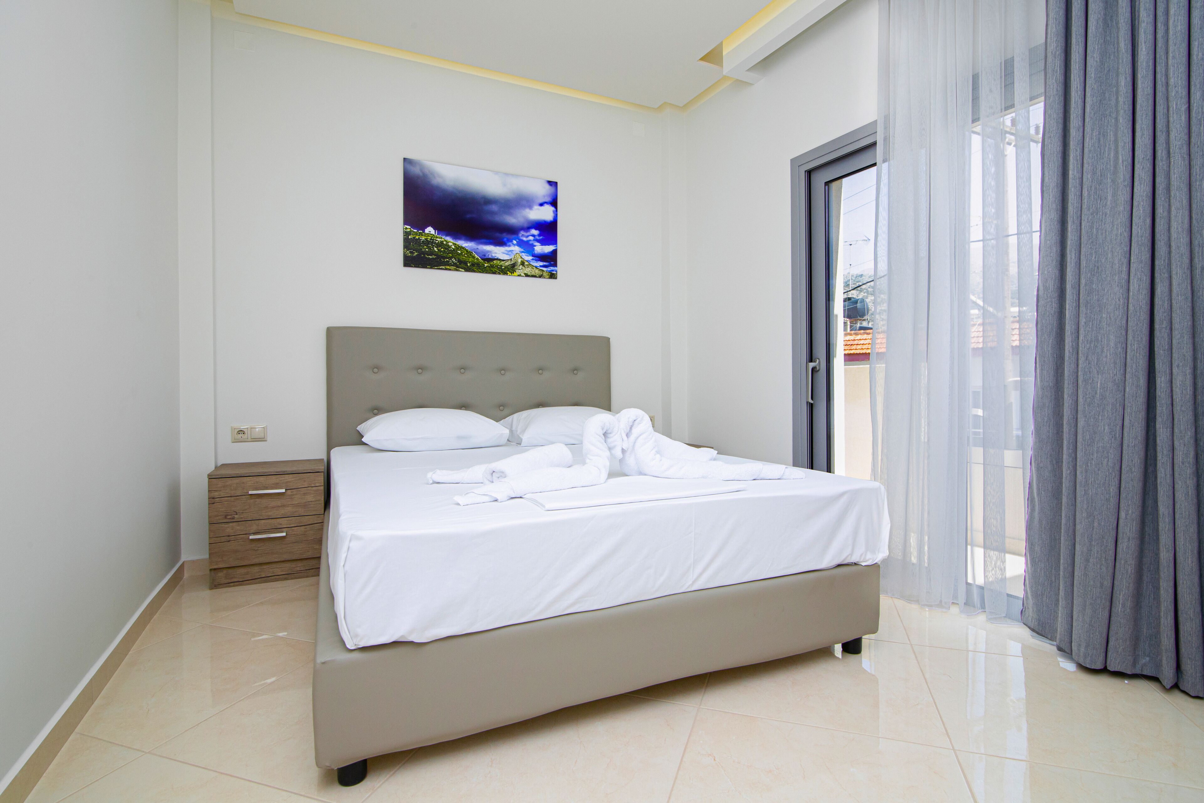 A neatly made bed with white linens, a grey headboard, and a small bedside table with drawers.