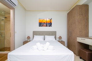 A neatly made bed with white linens and rolled towels, a wooden wardrobe, a bedside table with a lamp, and a painting of a sunset on the wall. - Amara Suites and Studios (Hersonissos)