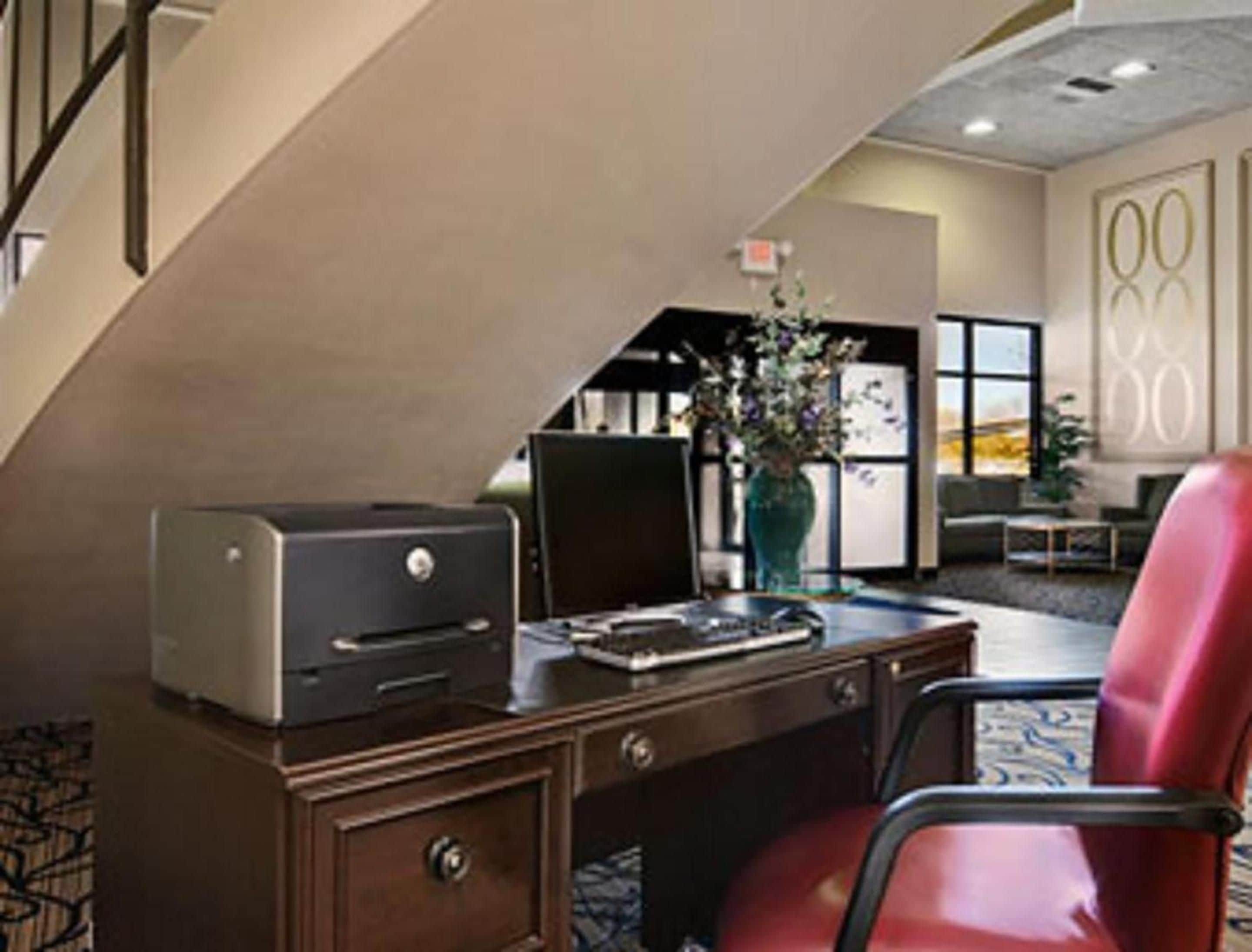 A hotel room with a desk, a printer, a vase with flowers, and a red chair.