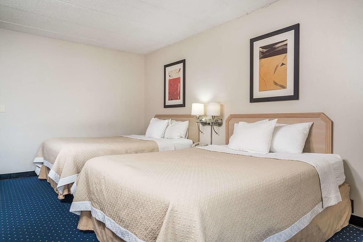Ein Hotelzimmer mit zwei Betten, jedes mit einer beigen Tagesdecke und weißen Kissen. An beiden Seiten der Betten befinden sich jeweils zwei Wandlampen.