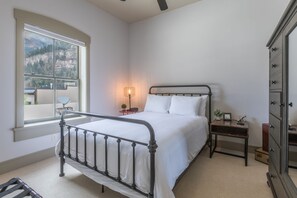 A bedroom with a metal bed frame, a bedside table with a lamp, a dresser, and a window with a view of mountains.