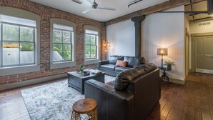 A spacious living room with a brick wall, large windows, a leather sofa, a coffee table, and a floor lamp. - Contemporary Cozy Loft | Heart of Downtown | Air Conditioning (Ouray)