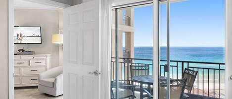 A balcony with a view of the ocean, a dining table with chairs, and a white door leading to a room with a TV and a dresser.