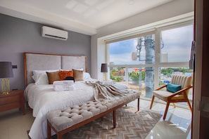 Une chambre moderne avec un grand lit, une vue sur la ville grâce à de grandes fenêtres et un balcon avec un fauteuil.
