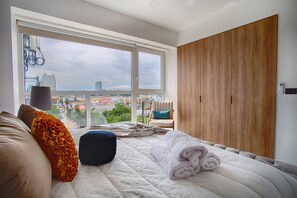 A bedroom with a large window, a bed with a white duvet, a wooden wardrobe, and a city view.