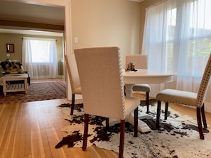 A dining area with a round table and four chairs, a cowhide rug, and a view of a garden through sheer curtains. - The Buckaroo- walk to historic downtown Cody, dog friendly! (Cody)