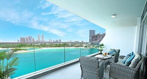 A balcony with wicker furniture, a table, and a view of a city skyline and water body. - LUX - Opulent Island Suite 6 (Dubai)