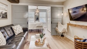 Un salon moderne avec un canapé en cuir, une table basse agrémentée d’objets décoratifs, un téléviseur fixé au mur et une vue sur la chambre à coucher par une porte.