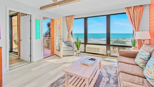 Uma sala de estar espaçosa com uma janela grande voltada para a praia, um espaço de estar acolhedor e uma mesa de centro de madeira.