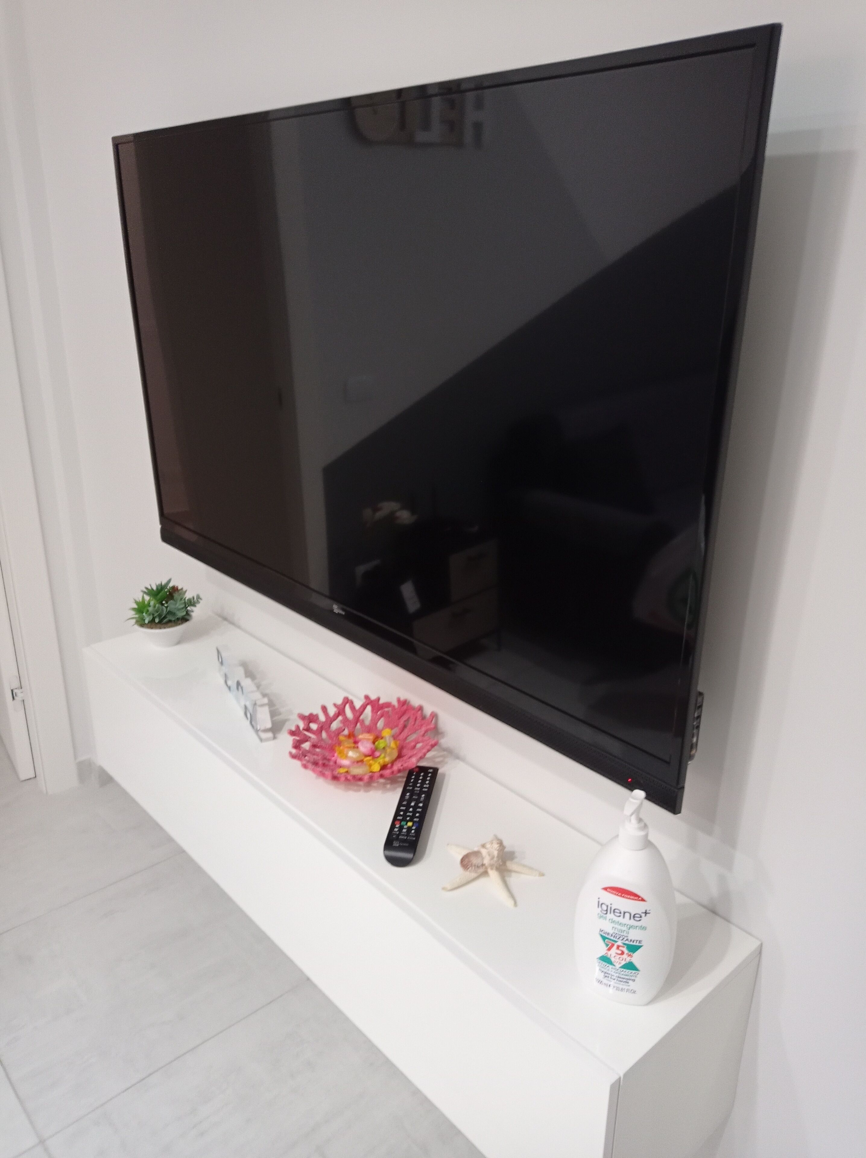 A flat-screen TV mounted on a white console with a remote, a decorative coral, a starfish, and a bottle of hygiene product.