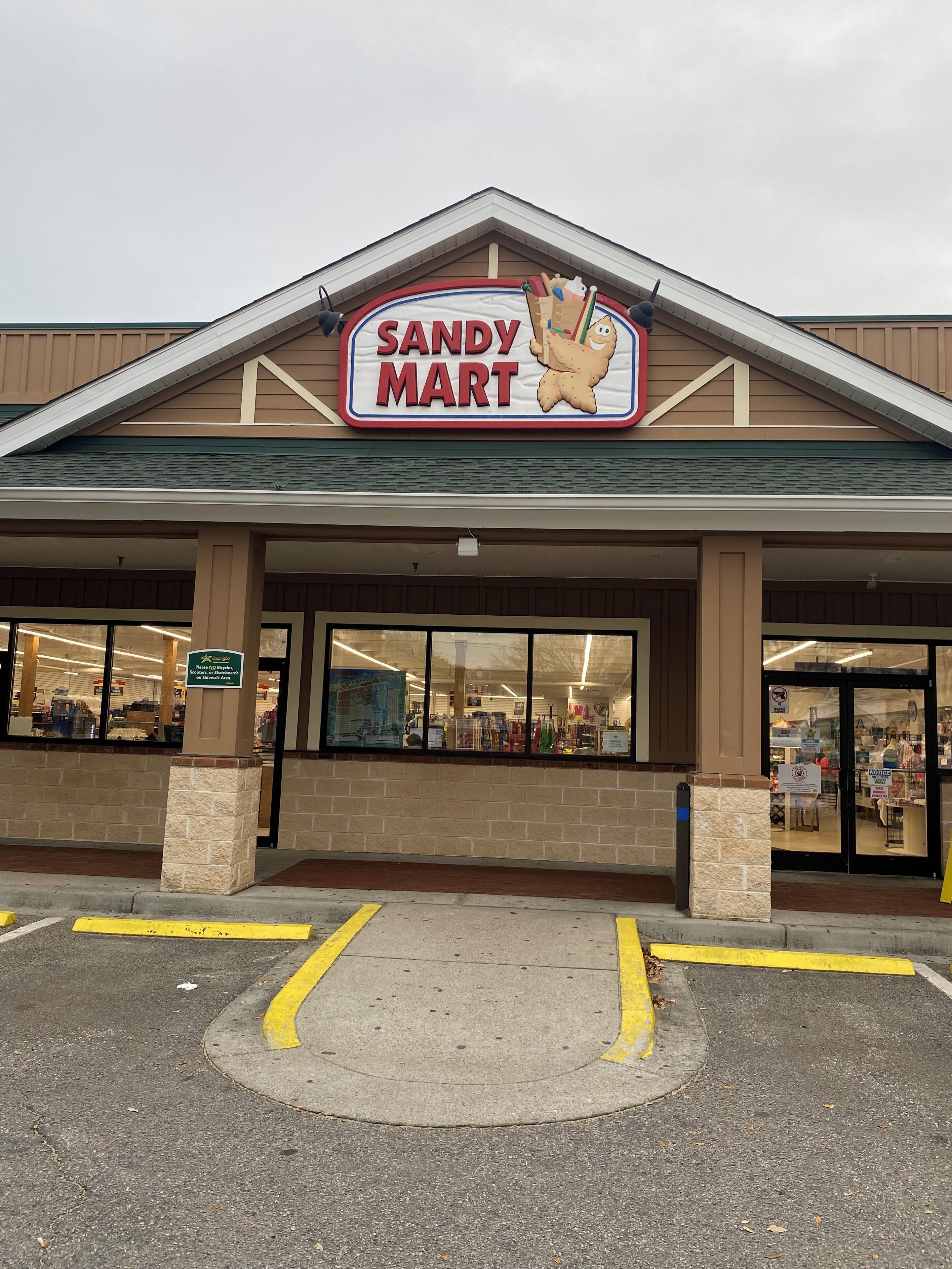 L’extérieur d’un magasin Sandy Mart, avec une entrée bien visible, un parking avec des places délimitées et une façade en briques.