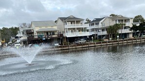 A waterfront view with a wooden post and rope barrier, a fountain, and residential buildings in the background. - Book now for the SUMMER before it’s gone @ Ocean Lakes Family Campground!! (Myrtle Beach)