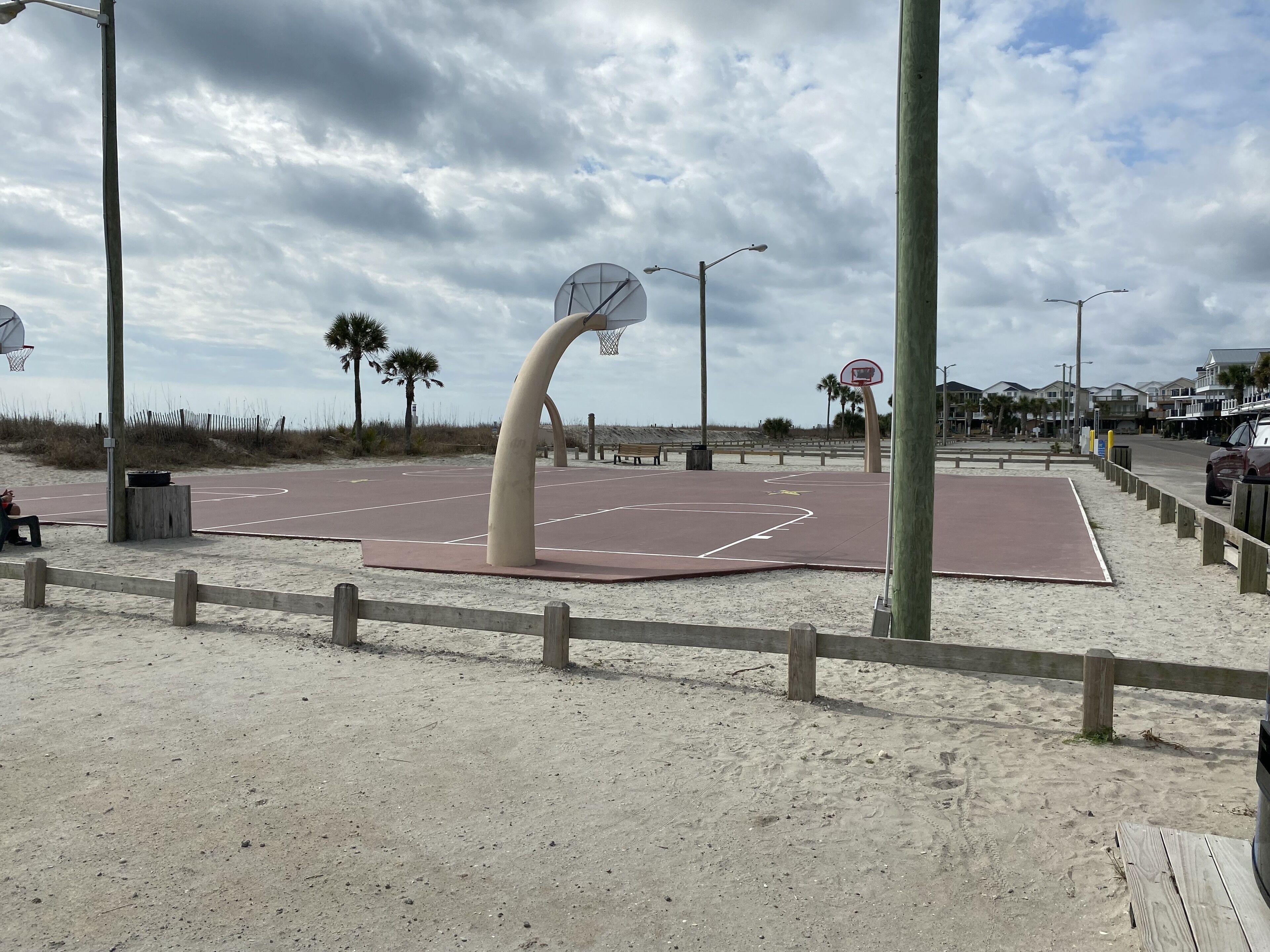 Un terrain de basket-ball aménagé sur la plage, doté d’un panier au design original, avec une structure incurvée, et entouré de sable et d’une clôture en bois.