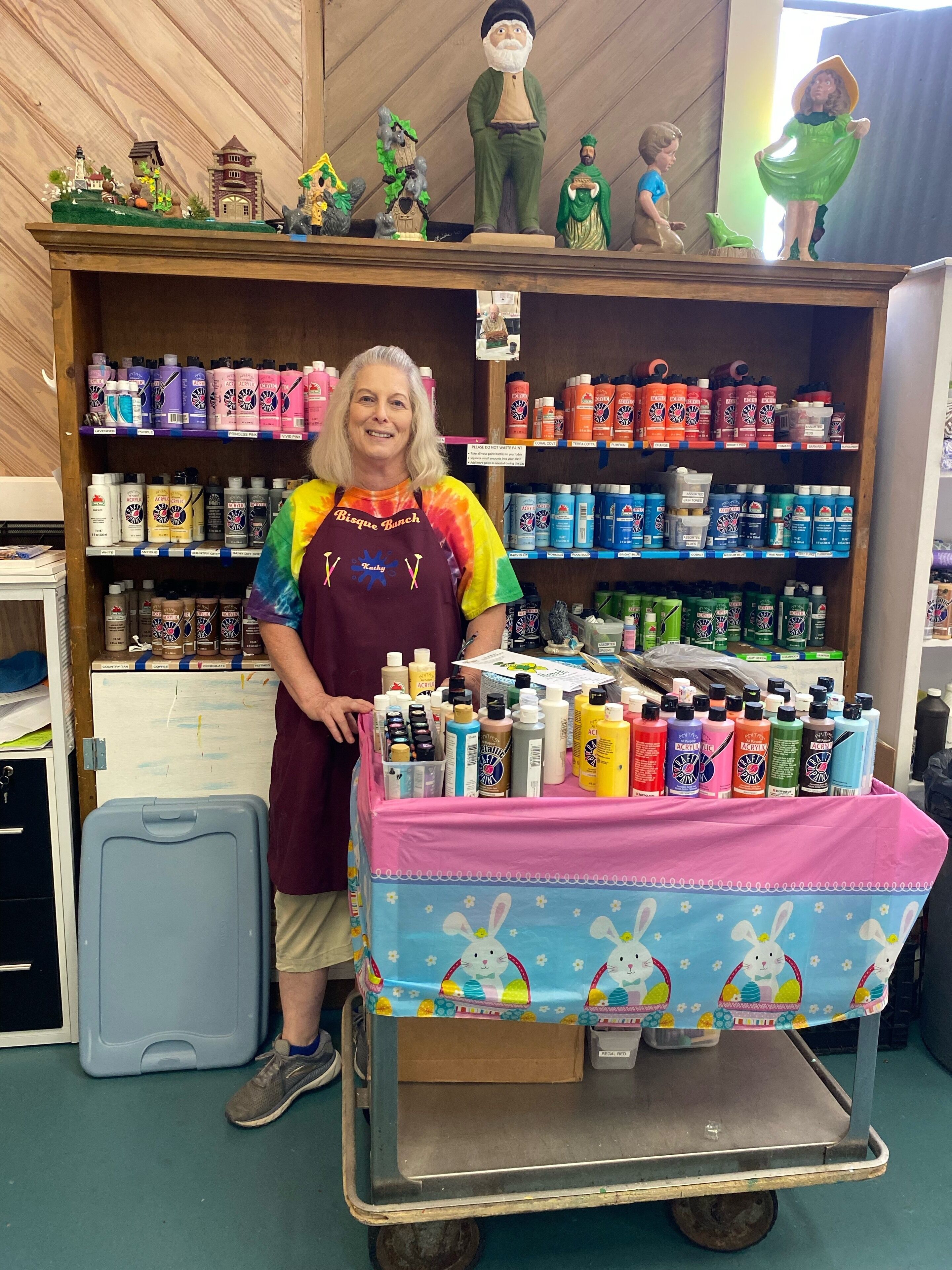 A person standing in a room with shelves stocked with various products, a cart with items on a table, and a small display of figurines.