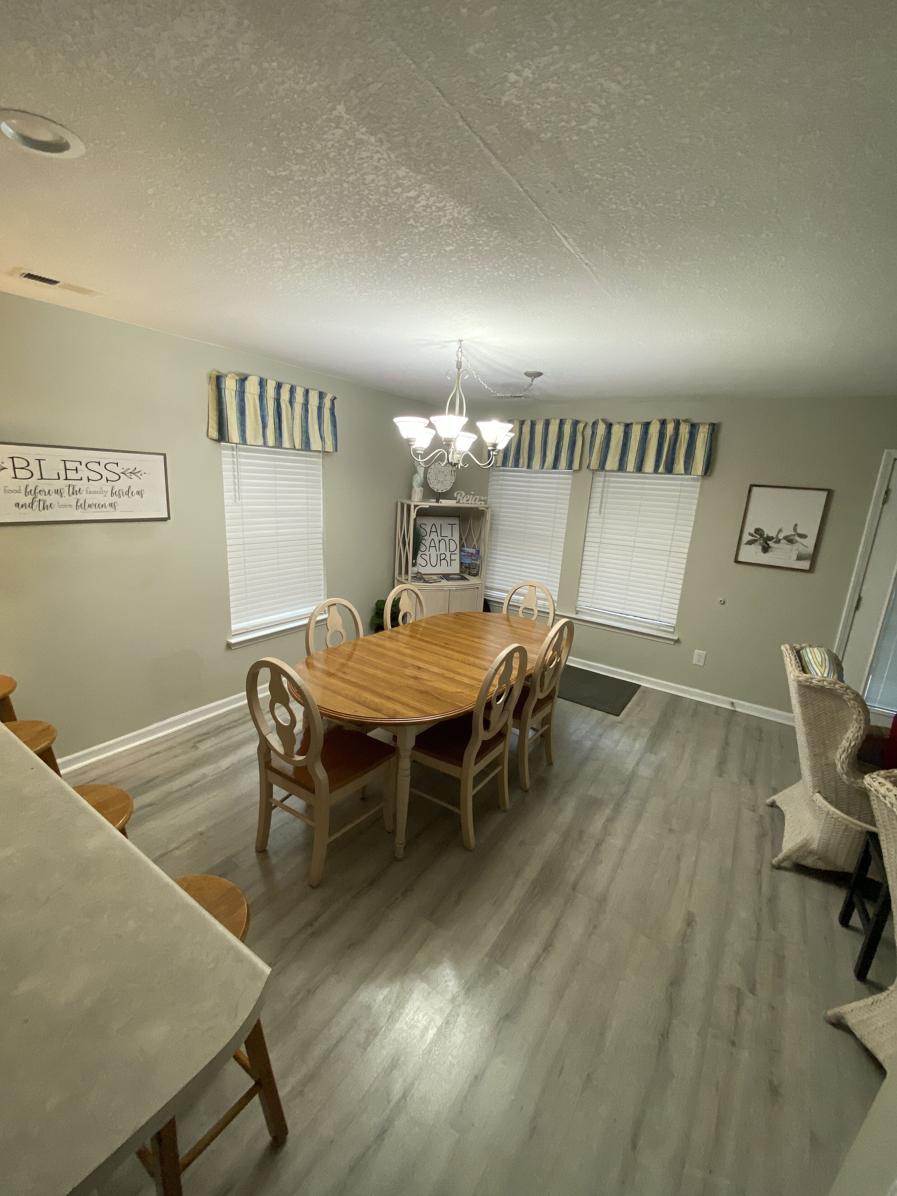 A dining room with a wooden table and chairs, a chandelier, and a sign that reads "Bless."