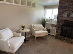 Uma sala de estar com dois poltronas brancas, uma mesa redonda pequena com um vaso de flores azul, um armário branco com prateleiras e uma lareira de pedra.