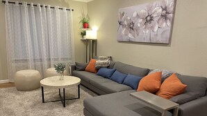 Un salon avec un canapé gris, un pouf et une table basse ronde. Un tableau est accroché au mur et une plante en pot est posée sur une étagère.