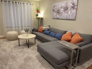 Sala de estar com sofá cinzento, banquinho e mesa de centro redonda. Há um quadro na parede e uma planta em vaso numa prateleira.