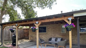 Un patio couvert avec un mur en briques, des meubles d’extérieur, des jardinières suspendues et un barbecue.