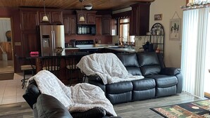 A living room with a black leather sofa, a kitchen in the background, and a wooden ceiling. - The Musky Cabin on the Big Eau Pleine Reservoir! (Auburndale)