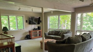 A living room with a sofa, a coffee table, a bookshelf, a television, and large windows with a view of trees. - Home in the woods - Big, Acreage, Nature Oasis, King Bed, Giant Windows (Monson)