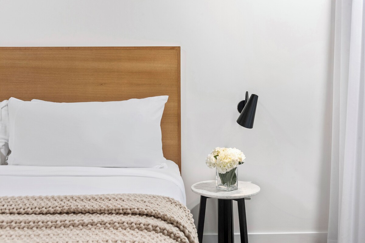 A bedroom with a wooden headboard, white bedding, a knitted throw, a small round table with a vase of flowers, and a black wall-mounted lamp.