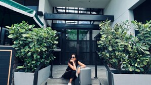 A person sitting on outdoor steps with a suitcase, surrounded by greenery and a modern building entrance. - UPSUN HOTEL (Miami Beach)