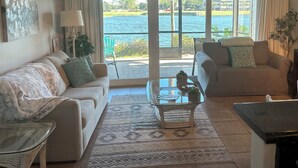A living room with two sofas, a coffee table, and a ceiling fan. There is a large window with a view of a body of water and greenery. - LAKEFRONT~BEACH~GOLF~POOLS~TENNIS~PET FRIENDLY (Miramar Beach)