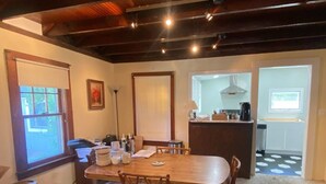 A dining area with a wooden table and chairs, a kitchen in the background, and a view through an open door. - Crescent Beach Cottage - Vacation or Academic Rental (Niantic)