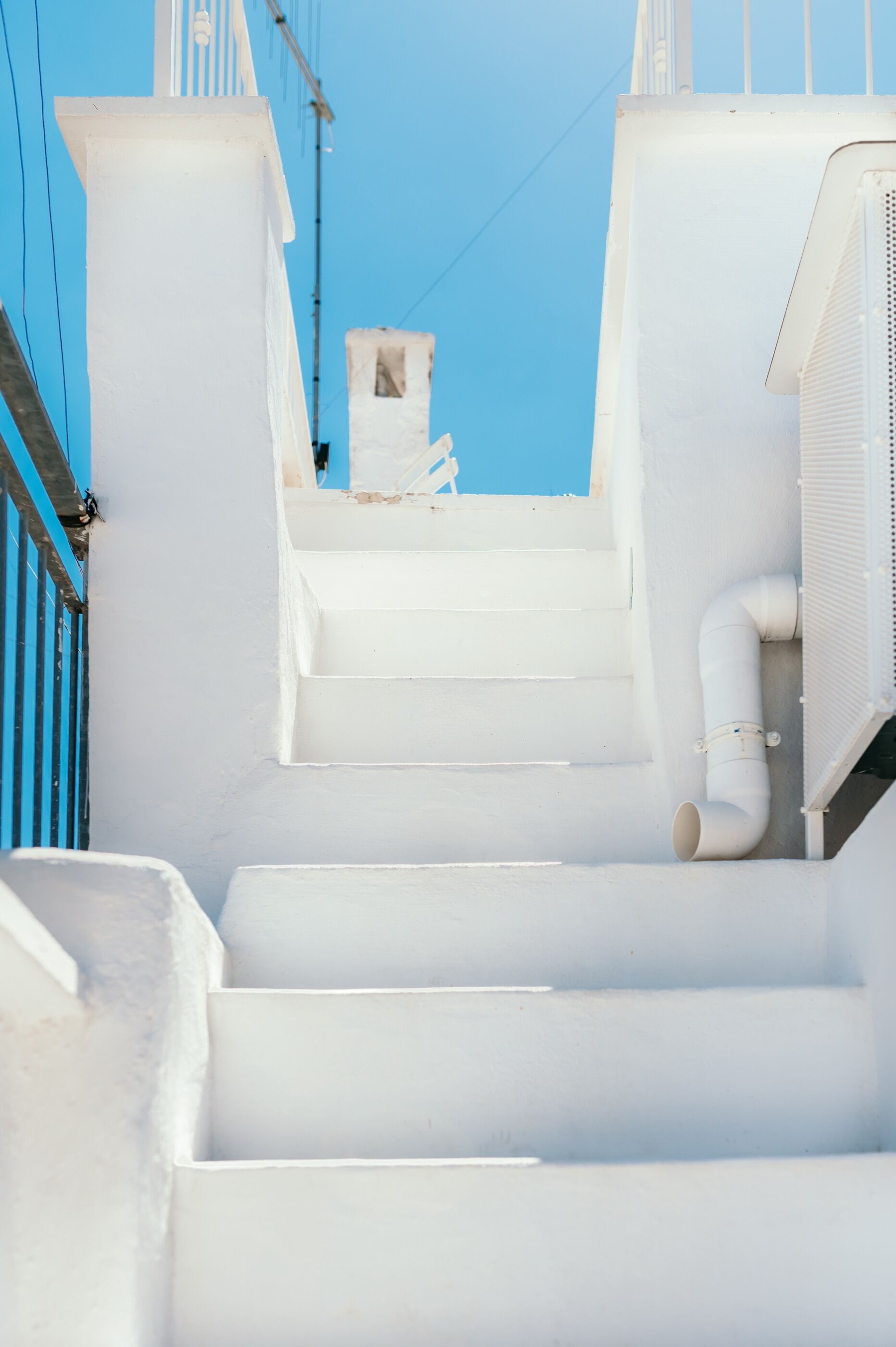Una scala bianca conduce a una terrazza sul tetto, con un edificio bianco e un cielo azzurro sullo sfondo.