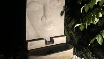 A stone sculpture of a face with a serene expression, set on a pedestal in a garden with pebbles and greenery.