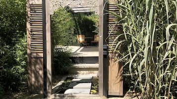 Un arc en bois orné de sculptures complexes, qui mène à un sentier de pierre bordé d’une végétation luxuriante et de plantes de bambou.