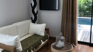 Un salon moderne avec un canapé blanc, une table basse ronde en bois, un téléviseur fixé au mur et une vue sur une terrasse en bois et la verdure environnante.