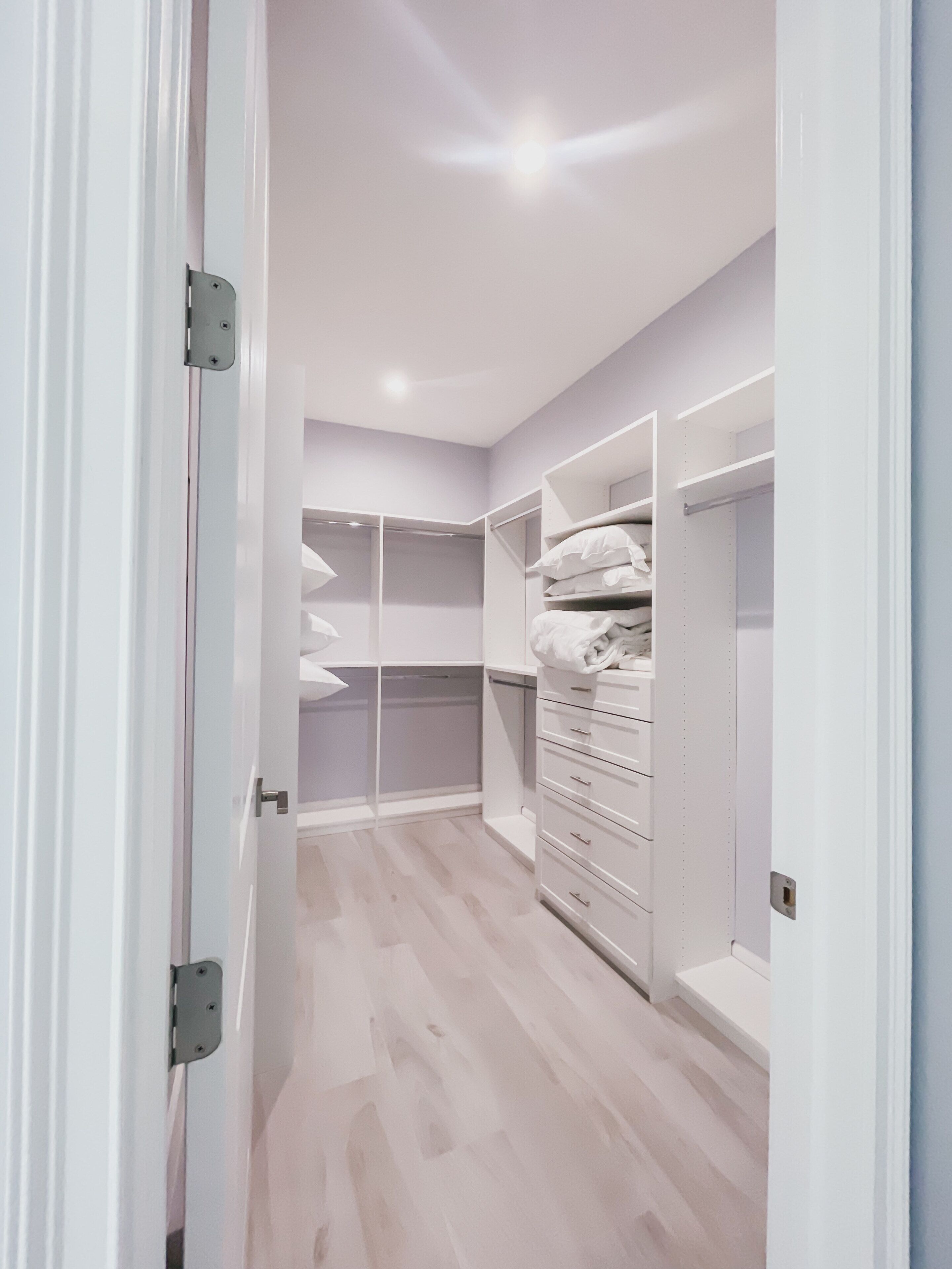 A walk-in closet with open shelving, drawers, and hanging space, featuring light wood flooring and white walls.