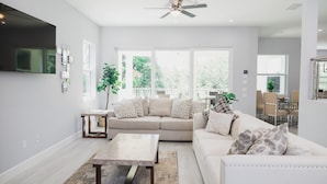 A spacious living room with a large window, a ceiling fan, and a dining area visible in the background. - Great reviews,Brand new,waterfront,heated pool, hot tub & beach access -14 pple  (Bonita Springs)