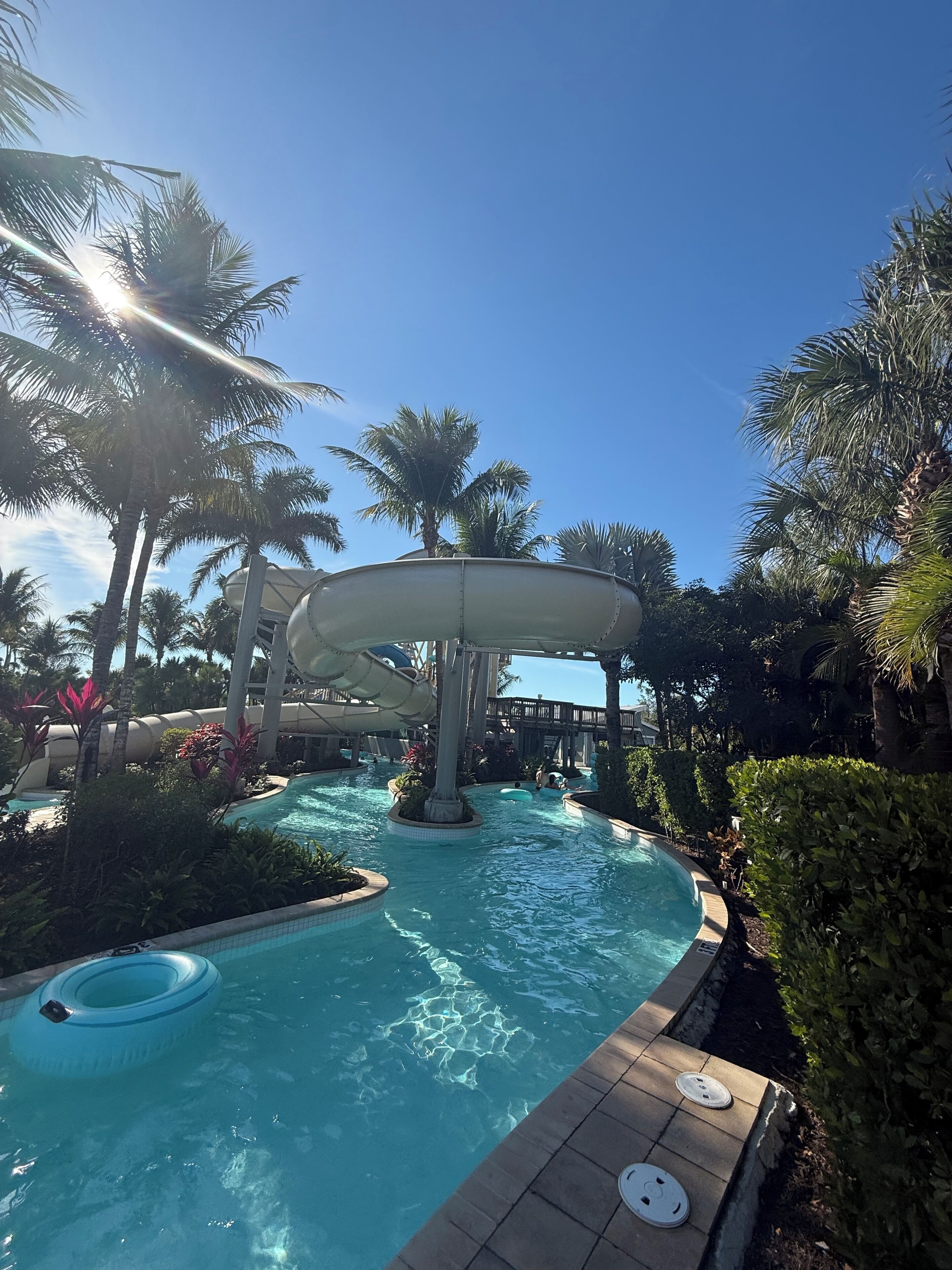 Un espace piscine doté d’un toboggan aquatique, de palmiers et surplombé d’un ciel d’un bleu éclatant.