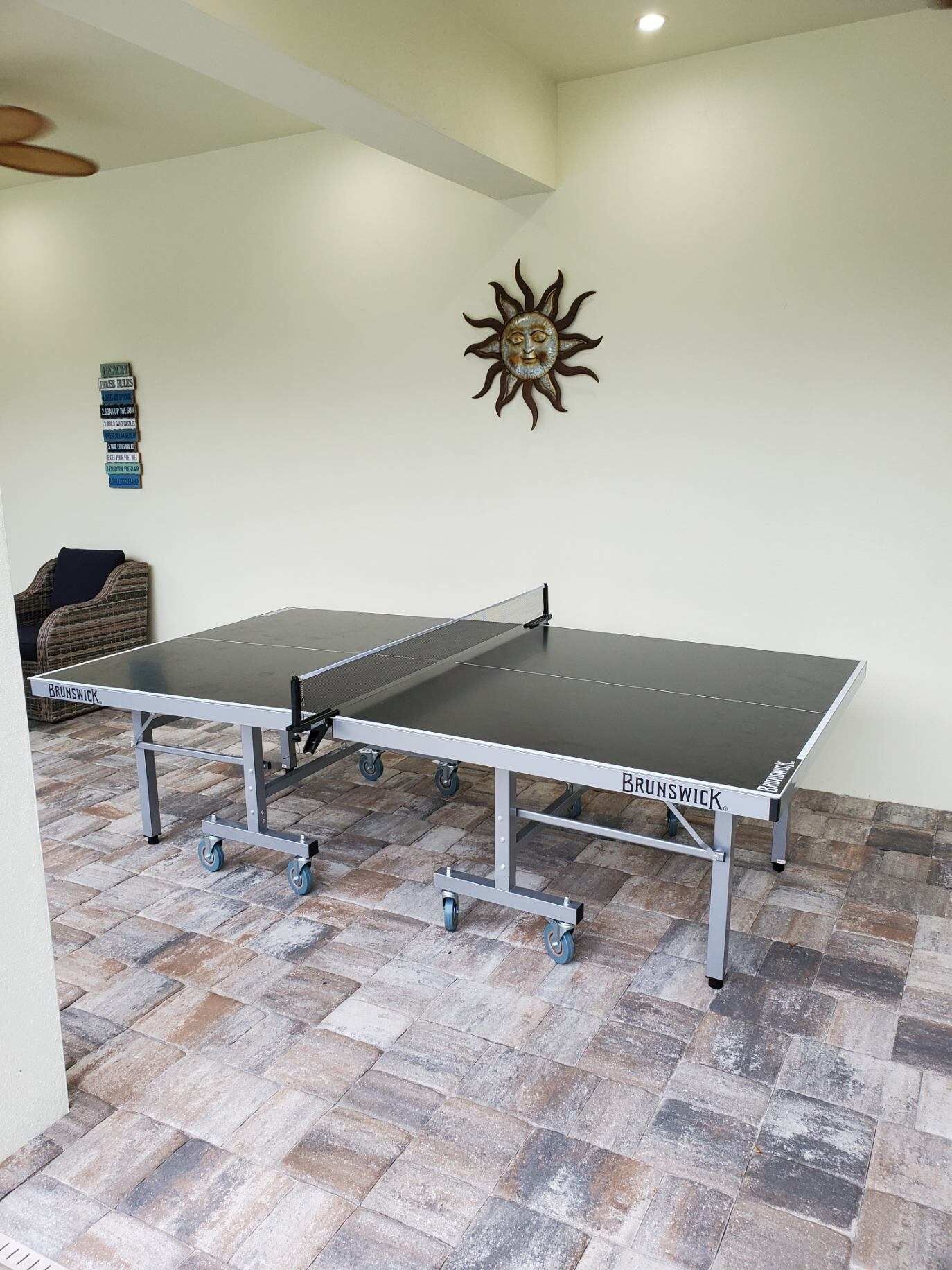 A ping pong table with wheels, labeled 'BRUNSWICK', in a room with a stone-tiled floor and a decorative sun wall art.