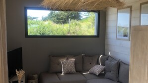 Ein modernes Wohnzimmer mit einer grauen Eckcouch, einem gewebten Teppich, einem großen Fenster mit Blick auf Grünflächen und einer Deckenleuchte.