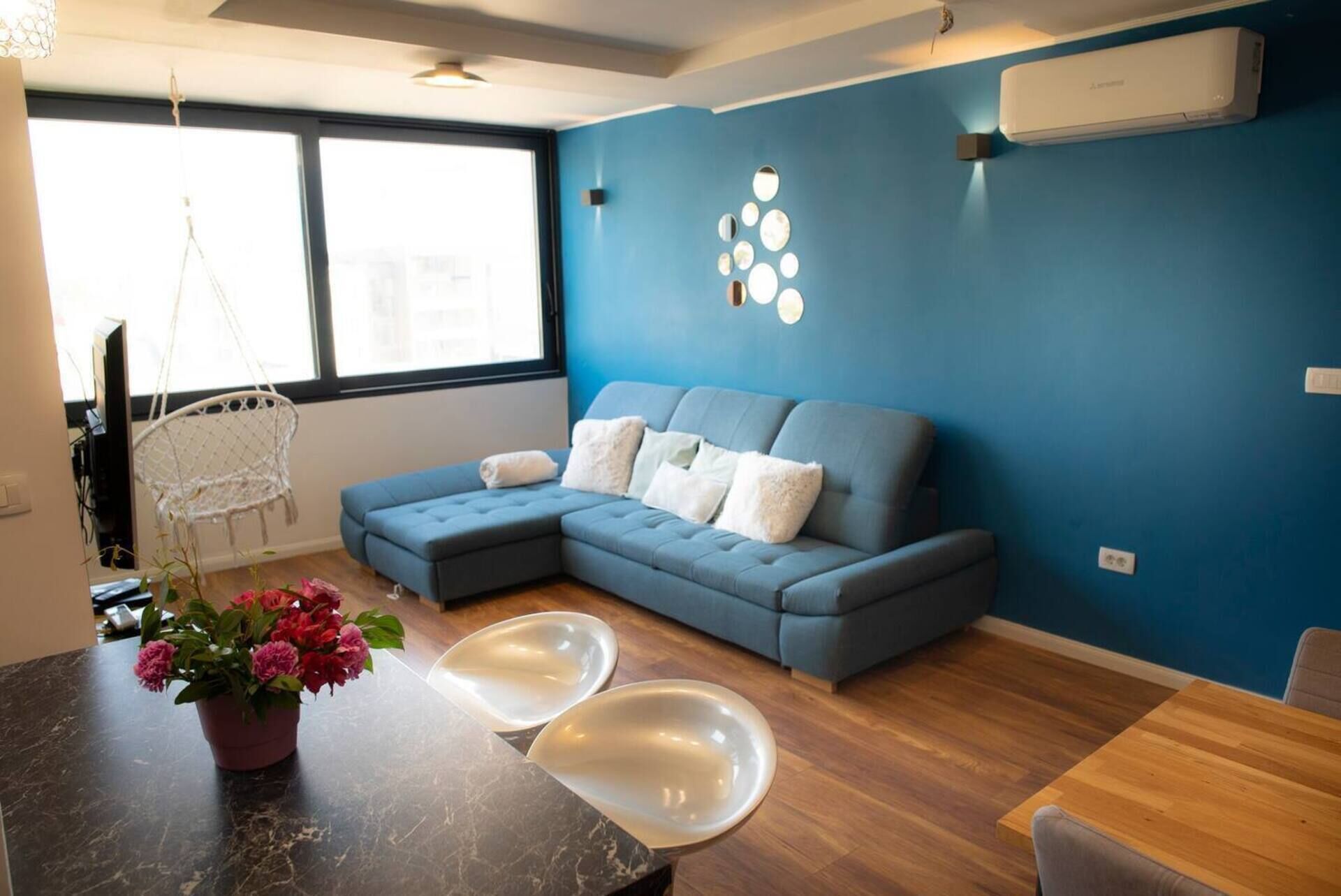 A modern living room with a sectional sofa, a hanging chair, a dining table with two chairs, and a decorative wall with circular mirrors.