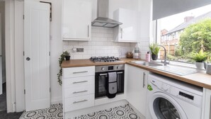 Cocina compacta con armarios blancos, estufa a gas, horno empotrado, lavadora y ventana con vistas al verdor.