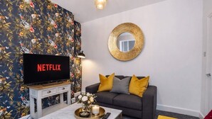 Una sala de estar con una televisión mostrando 'NETFLIX', un sofá con cojines amarillos, un espejo redondo en la pared y una mesita con una vela y flores.