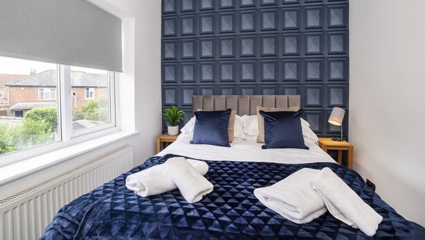 Una cama bien hecha con una colcha estampada oscura, dos almohadas y dos toallas dobladas.