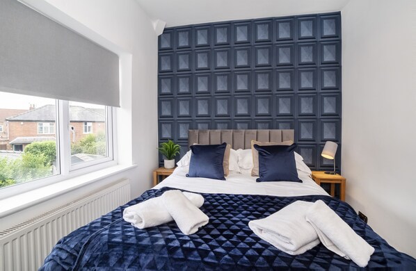 A neatly made bed with a dark patterned bedspread, two pillows, and two folded towels.