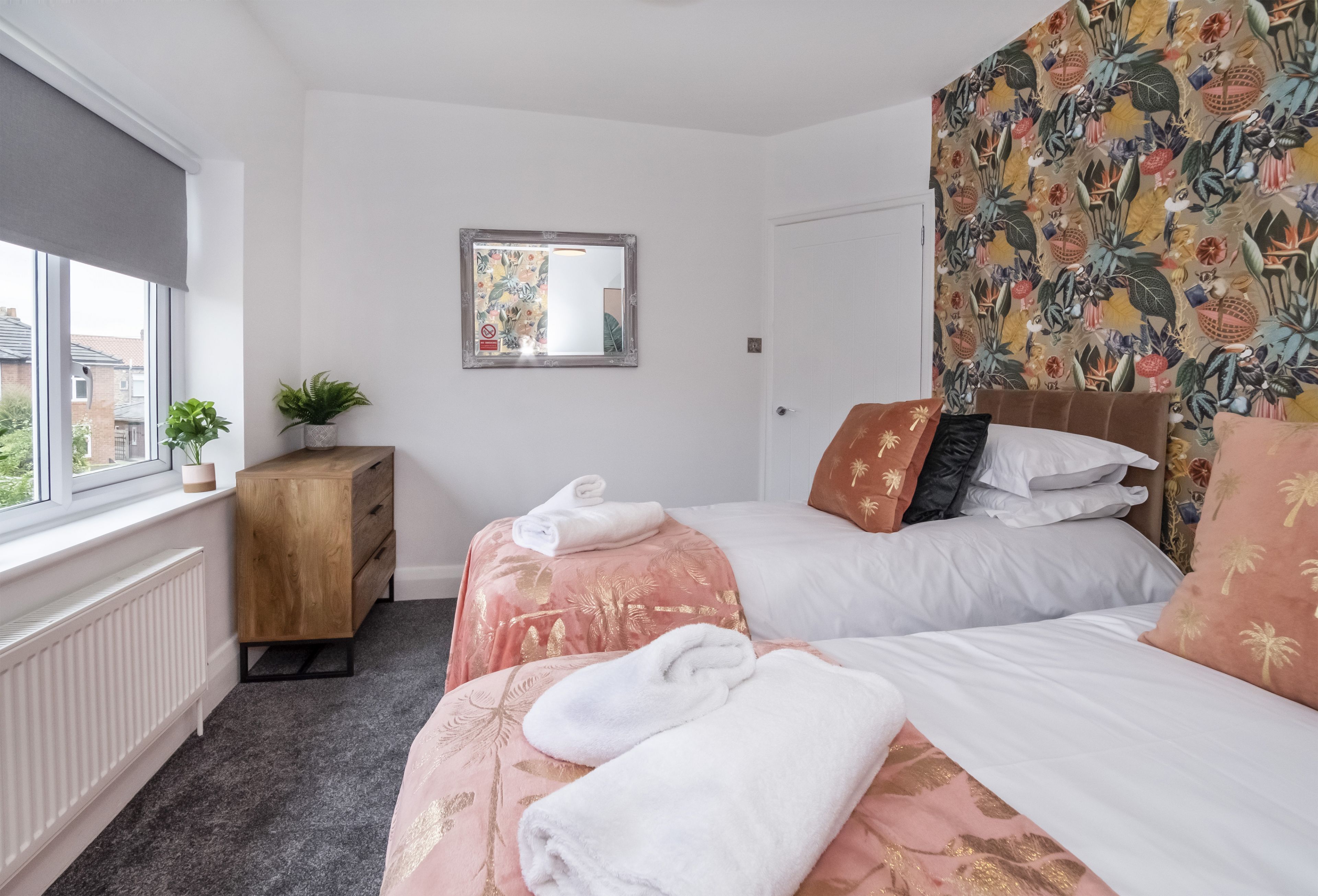 Quarto com duas camas, cômoda de madeira, radiador, janela com vista e espelho na parede.